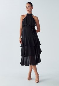 Musta olkaimeton mekko, kerroksellinen röyhelöinen muotoilu, virtaava läpikuultava kangas, solmittu vyötärön yksityiskohta ja nilkkapituinen leikkaus. Siinä on hillitty kiilto.