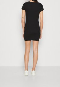 Femme debout, de dos, portant une robe mini noire à manches courtes ajustée et des baskets blanches, sur un fond clair uniforme.