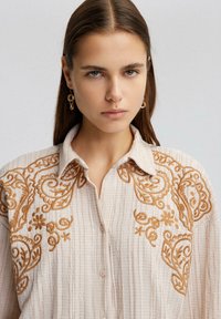 Light beige shirt featuring intricate rust-colored embroidery with floral and swirl patterns, collared design, and button front.