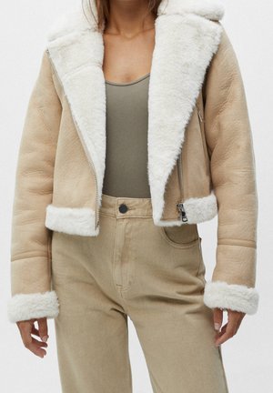 Vrouw draagt een beige suède jas met witte schapenvacht voering en manchetten, gecombineerd met beige broek met hoge taille en een olijfgroen topje.