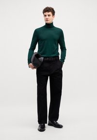 Grüner Rollkragenpullover, schwarze plissierte Hose und schwarze Lederschuhe. Model hält eine schwarze Tasche mit minimalistischem und schlankem Design.