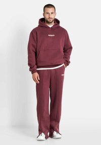 Bordeauxfarbener Hoodie mit Fronttasche, weißem Markenlogo, passende Hose mit seitlichen Schlitzen und weißen Schuhen. Weicher Stoff, entspannte Passform.