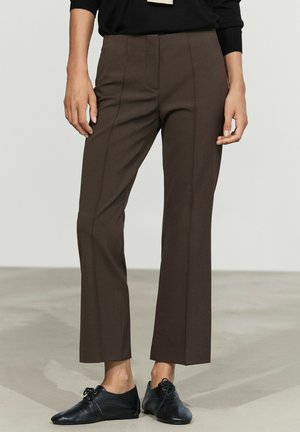 Femme portant un pantalon marron droit et des chaussures plates noires à lacets, debout sur un sol lisse gris clair.
