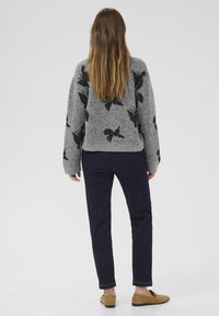 Pelēks adīts džemperis ar melniem lapu rakstiem, apvienots ar tumši ziliem džinsiem un gaiši brūniem plakanajiem apaviem. Redzami gari, taisni mati.