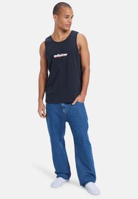 Tricou din bumbac bleumarin, cu guler rotund și logo-ul Quiksilver. Asortat cu blugi slăbiti din denim albastru și pantofi albi slip-on.