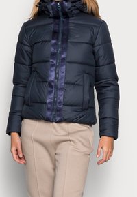 Marinblå pufferjacka med ett quiltat mönster, som har en glänsande mörkblå rand i mitten, en huva och fickor fram med dragkedja.