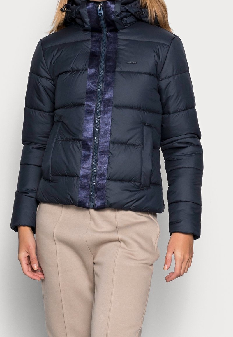 Marinblå pufferjacka med ett quiltat mönster, som har en glänsande mörkblå rand i mitten, en huva och fickor fram med dragkedja.