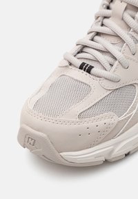 Baskets de sport beige clair avec des panneaux en maille, semelle en caoutchouc, languette rembourrée et système de laçage avec des accents noirs et un logo.