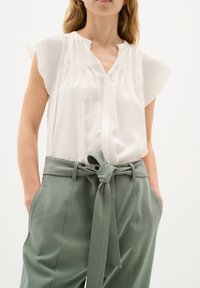 Blusa bianca a maniche corte con scollo a V e dettagli plissettati, abbinata a pantaloni verde oliva a vita alta con cintura annodata e tasche.