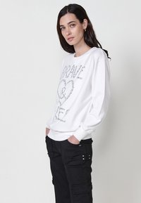 Felpa bianca con la scritta “BRAVE” in decorazioni argentate, scollo rotondo, spalle abbassate e maniche lunghe, abbinata a pantaloni cargo neri.