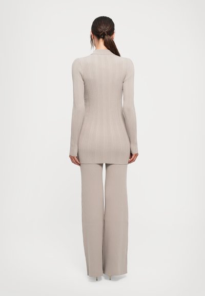 Woman with brown ponytail wearing beige ribbed long-sleeve top and matching wide-leg pants, standing against white background, back view.
