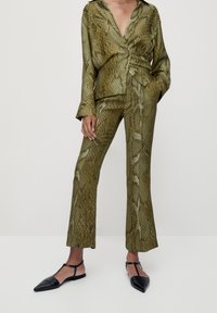 Ensemble assorti en imprimé serpent vert olive comprenant une chemise enveloppante avec détail à nouer et un pantalon évasé, associé à des ballerines noires à bouts pointus.