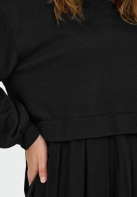 Maglione nero corto realizzato in tessuto morbido, con maniche lunghe e vita aderente, abbinato a una gonna nera plissettata.