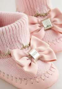 Rozā bērnu zābaciņi ar adītu augšdaļu, dekorēti ar lielu auduma loku un sudraba akcentu. Mīksta tekstūra ar izšūšanas detaļām gar zoles malu.