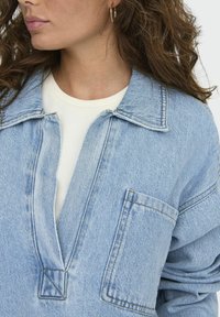 Camisa de denim en azul claro, con un diseño sin cuello, bolsillo frontal y un corte holgado. Tejido texturizado con detalles de costura visibles.