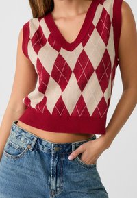 Červený a krémový svetr bez rukávů ve vzoru argyle s výstřihem do V a žebrovaným lemem, kombinovaný s džíny z modré denimu s vysokým pasem.