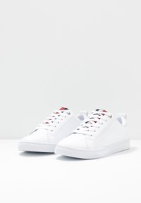 Baskets blanches avec un dessus synthétique lisse, bout rond et semelle en caoutchouc plate. Présente des accents rouges et marins sur la languette.