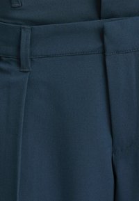 Mörkgröna byxor i mjukt tyg. Har en platt midjeband med bälteshällor och en prydligt sydd veck framtill.