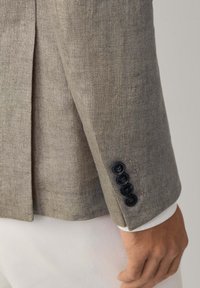 Manche d'un blazer en lin beige avec quatre boutons noirs, porté sur une chemise blanche et un pantalon clair, la main reposant sur le côté.