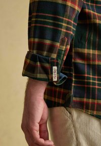 Flaneļa krekls ar zaļu, tumši zilu, sarkanu un dzeltenu svītru rakstu. Iekļauts ar piedurkni, uz kura ir zīmola etiķete, un teksturētu audumu.