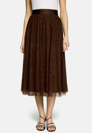 Femme portant une jupe midi plissée marron avec un voile transparent, associée à des sandales noires à brides et un haut blanc sans manches.