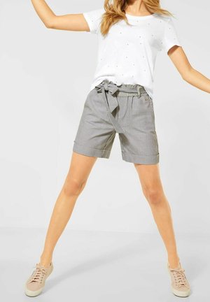 Woman wearing white short-sleeve shirt with small dots and high-waisted striped shorts tied with a bow, standing with legs apart.