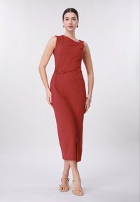 Γυναίκα που φοράει ένα αμάνικο μεσαίο φόρεμα σκουρόχρωμου κόκκινου με λεπτομέρεια αγκράφας στο πλάι, στέκεται με τα χέρια χαλαρά, με μπεζ σανδάλια με λουριά και απλά κοσμήματα.