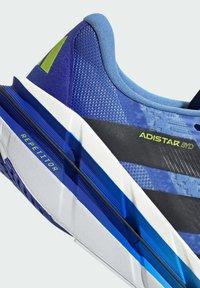 Blue athletic shoe with textured mesh upper, contrasting black and white midsole, and green accents. Features bold branding and dynamic lines.