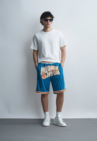 Fiatal férfi fekete napszemüveget visel, fehér pólót, kék "Billionaire Boys Club" rövidnadrágot, fehér zoknit és fehér sportcipőt, egy sima fal előtt áll.