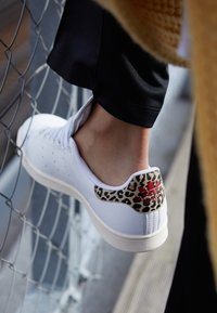 Weiße Ledersneaker mit perforierten Details, Akzent in Leopardenmuster an der Ferse und rotem Branding. Texturierte Sohle. Positioniert vor einem Zaun.