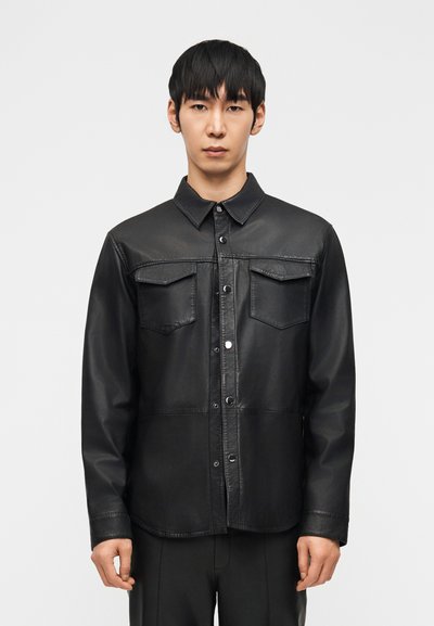Jeune homme aux cheveux noirs raides portant une chemise noire en similicuir boutonnée avec des poches poitrine, sur un fond clair uni.