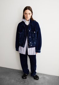 Giacca di denim in corda blu scuro, abbinata a una camicia a righe con bottoni e pantaloni coordinati blu scuro. Scarpe nere testurizzate completano il look.