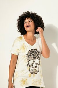 T-shirt à col en V crème avec éclaboussures dorées. Présente un graphique de crâne en imprimé léopard. Coupe décontractée avec des manches courtes et des fentes sur les côtés.