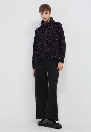 Schwarzer strukturierten Pullover mit bunten Sprenkeln, mit hohem Kragen. Kombiniert mit schwarzen weit geschnittenen Hosen mit Seitenschaltern und schwarzem Schuhwerk.