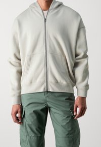 Felpa con zip color avorio e cappuccio, con tasche frontali e polsini a coste, indossata con pantaloni cargo verdi, realizzati in un tessuto testurizzato.