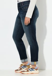 Tmavě modré džíny s úzkým střihem, ohrnutými lemy a zipovou kapsou. V kombinaci s vícebarevnými teniskami se šňůrkami.