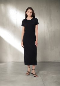 Črna maxi obleka s kratkimi rokavi iz mehke tkanine, s poudarjeno oprijeto silhueto. Oblečena s črnimi sandali s peto in trakovi.