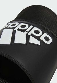 Černé skluzné sandály s širokým popruhem a bílým logem a výrazným grafickým designem. Hladký povrch s matným povrchem. Jednoduchý, otevřený tvar pro prsty.