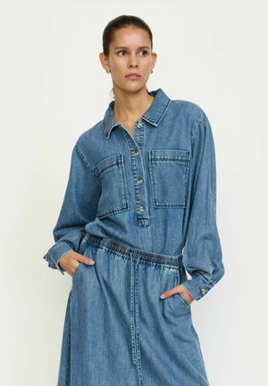 Das Jeanshemdkleid verfügt über eine Knopfleiste auf der Vorderseite, lange Ärmel, zwei Brusttaschen und einen elastischen Taillenbund. Die Farbe ist hellblau mit einer glatten Textur.