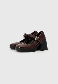 Des chaussures Mary Jane en cuir verni marron avec une tête arrondie, une sangle ajustable, une boucle en argent et un talon épais noir. Texture lisse.