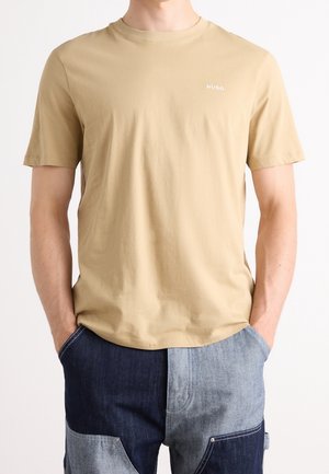 Beżowy T-shirt z krótkim rękawem z logo "HUGO" na piersi, noszony przez osobę z rękami w kieszeniach spodni z ciemno- i jasnoniebieskiego dżinsu typu patchwork.