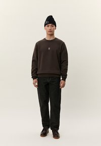 Joven con gorro negro, sudadera marrón con el logo "II", vaqueros oscuros y zapatos marrones, de pie frente a un fondo claro y liso.