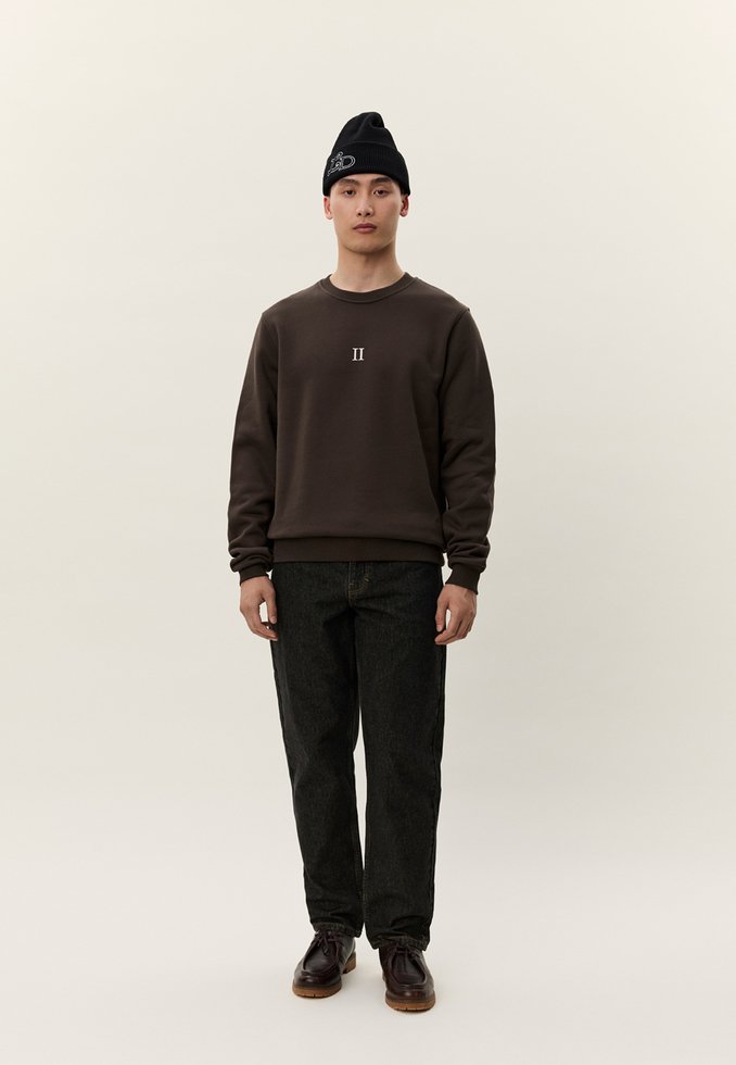 Joven con gorro negro, sudadera marrón con el logo "II", vaqueros oscuros y zapatos marrones, de pie frente a un fondo claro y liso.