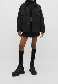 Sort oversized jakke med to forlommer, sort crop top, stribet sort plisseret nederdel, knæhøje sokker og kraftige sorte støvler.