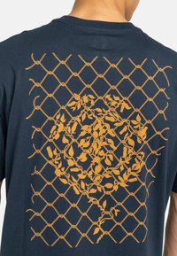 Marineblauw katoenen T-shirt met een groot oranje bloemen- en geometrisch hekpatroon op de achterkant, heeft een ronde halslijn en korte mouwen.