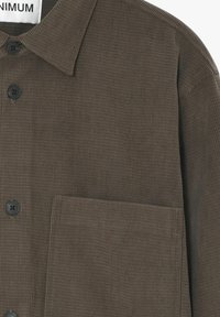 Hnedá kordurová košeľa s golierom na gombíky, prednou náprsní kapsou a čiernymi gombíkmi. Tkanina má ribovanú textúru.