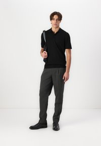 Crna majica polo s kratkim rukavima, u kombinaciji s tamno sivim prugastim hlačama. Model drži crnu torbu za rame i nosi crne cipele.