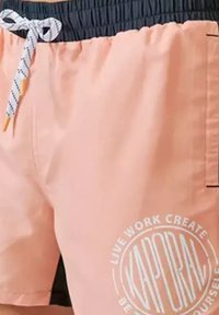 Shorts de bain pêche avec une ceinture élastique marine, un cordon de serrage blanc et un logo rond sur le devant avec des accents de texte et graphiques.