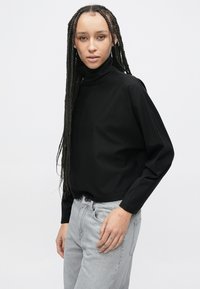 Černý top s dlouhým rukávem a vysokým límcem, vyrobený z hladkého materiálu, v kombinaci se světle šedými džínami s volnějším střihem.