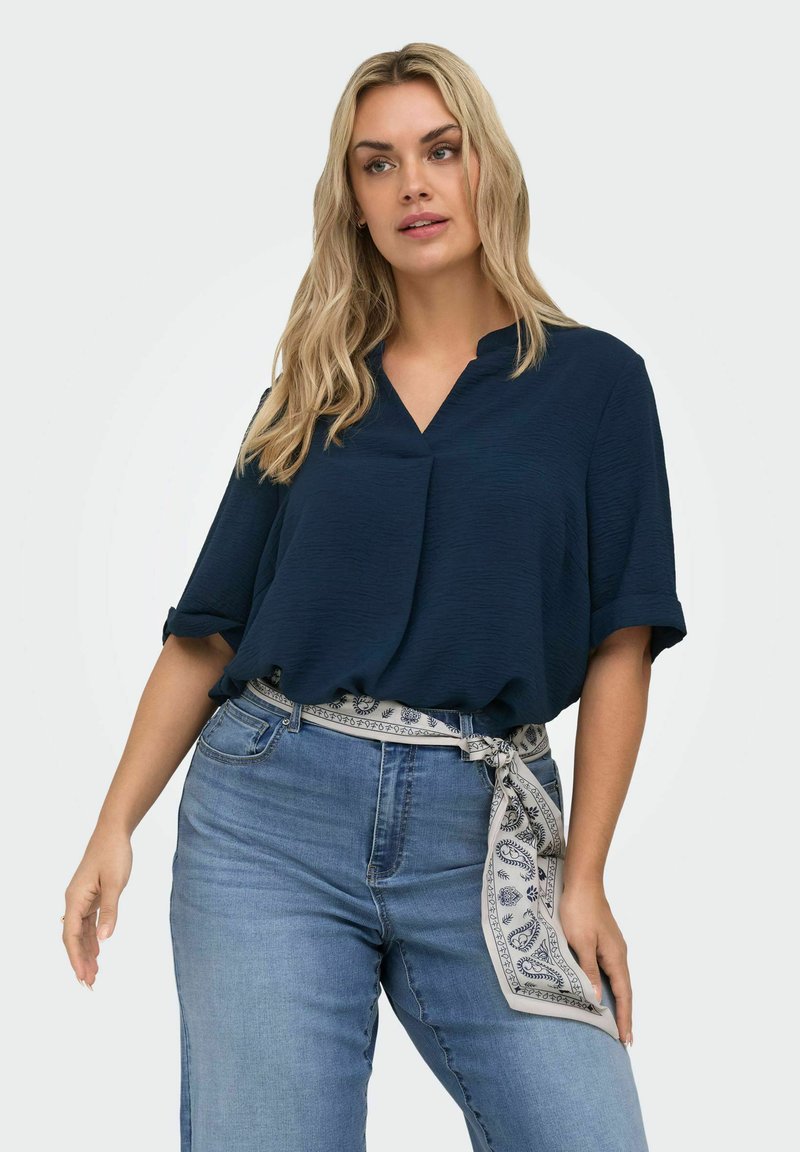 Žena oblečená v tmavomodrej blúzke s vyhrnutými rukávmi, svetlomodrých džínsoch a bielom šatkovom páse s vzorom previazanom v páse.